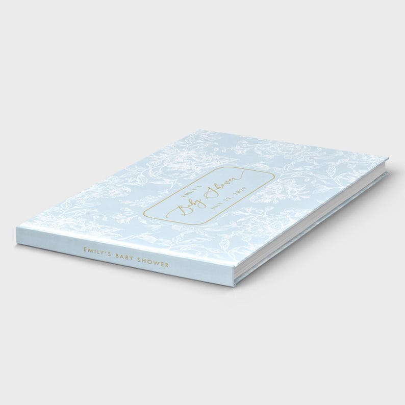 May include: A light blue baby shower guest book with a floral pattern. The book has gold lettering that reads "Emily's Baby Shower" and the date. The book is closed and lying on a white surface.