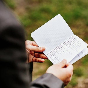 May include: A person holding a small, white notebook with lined paper. The notebook is open and the pages are filled with handwritten notes.