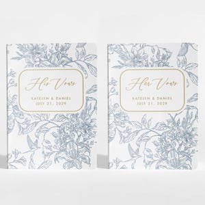 May include: Two wedding vow books with floral designs in blue and white. The text "Her Vows" is in gold script, with the names "Katelyn & Daniel" and the date "July 21, 2029" below. The books are side by side.
