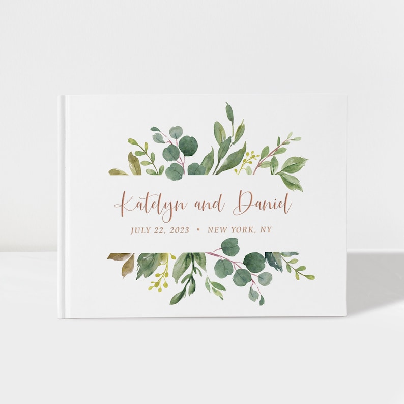 May include: White wedding guest book with a watercolor design of green leaves and branches. The text "Katelyn and Daniel" is written in a copper-colored font. The date "July 22, 2023" and location "New York, NY" are written below the names.