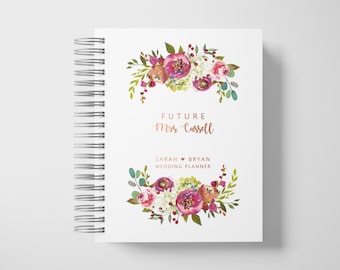 May include: White spiral-bound wedding planner with floral watercolour design. The cover features the text "FUTURE Mrs. Cassell" and "SARAH & BRYAN WEDDING PLANNER" with a floral arrangement in pink, purple, and green.