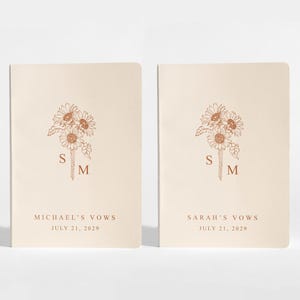 May include: Two cream-colored vow books with a sunflower illustration and initials. The text "MICHAEL'S VOWS JULY 21, 2029" and "SARAH'S VOWS JULY 21, 2029" are printed below the floral design. The books are for a wedding.