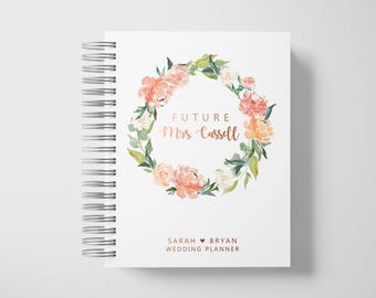 May include: A white wedding planner with a spiral binding. The cover features a floral wreath in shades of peach, pink, and green, encircling the words "FUTURE Mrs. Cassell." Below, it reads "SARAH ️ BRYAN WEDDING PLANNER."