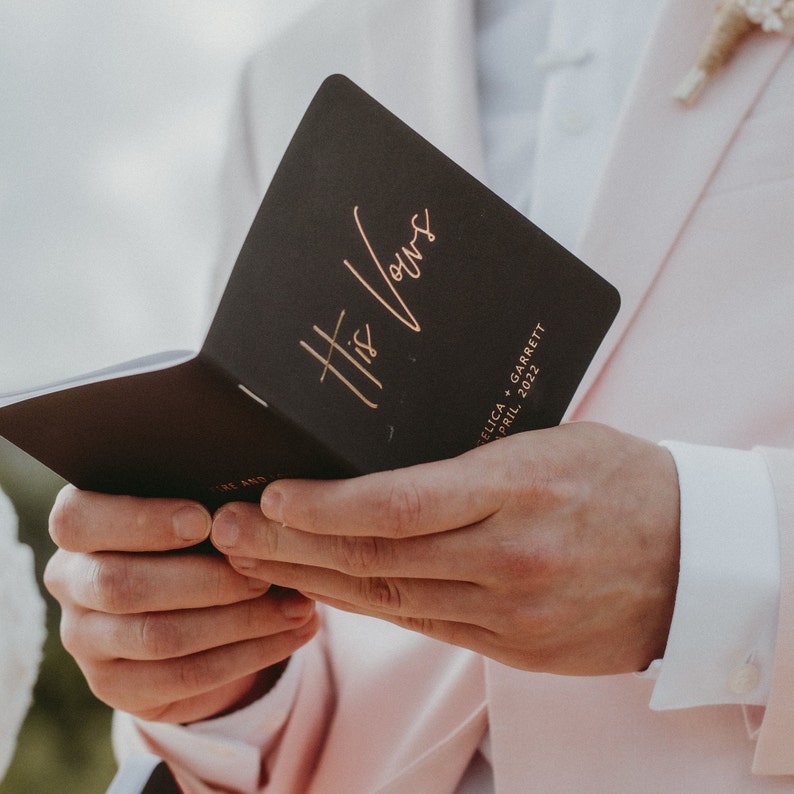 May include: A black booklet with gold lettering that reads "His Vows" and the text "Felica + Garrett April 2022".