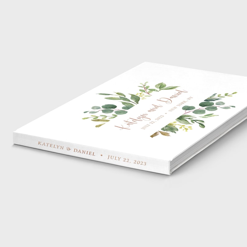 May include: A white wedding guest book with a leafy green watercolor design and the text "Katelyn and Daniel" in a copper-colored font. The date "July 22, 2023" is printed below the names.