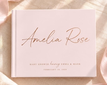 May include: A light pink hardcover guest book featuring the name Amelia Rose in elegant copper-colored script. Below, the text reads BABY SHOWER honoring EMMA & MARK and FEBRUARY 10, 2029. The book is surrounded by a soft, flowing pink ribbon.