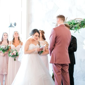 May include: A bride in a white wedding dress reads a letter during her wedding ceremony. She is standing in front of the groom, who is wearing a pink suit. The bride's bridesmaids are standing behind her, wearing blush-colored dresses.