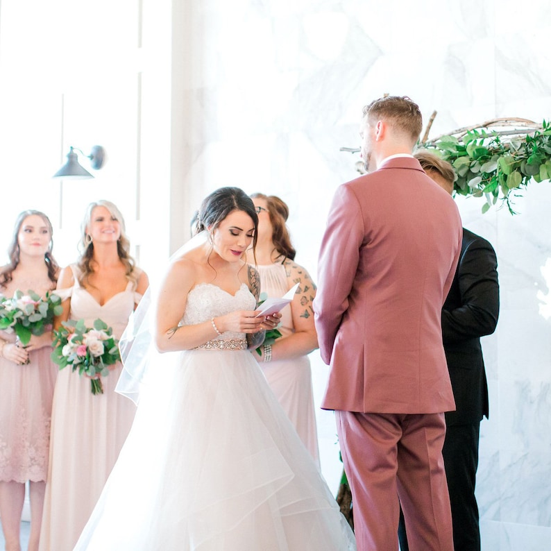 May include: A bride in a white wedding dress reads a letter during her wedding ceremony. She is standing in front of the groom, who is wearing a pink suit. The bride's bridesmaids are standing behind her, wearing blush-colored dresses.