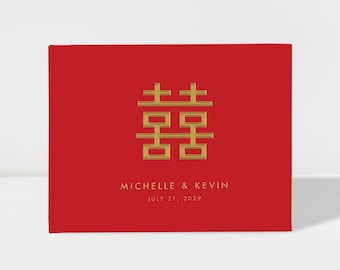 May include: A rectangular red book with a gold double happiness symbol centered on the front cover. Below the symbol, the text reads MICHELLE & KEVIN and JULY 21, 2029 in gold lettering. The cover has a smooth, matte finish and a clean design.