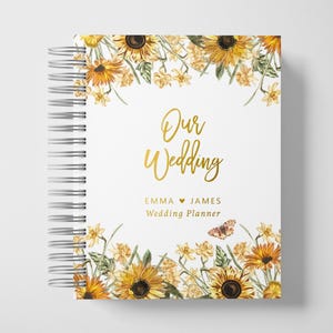 Puede incluir: Un planificador de bodas encuadernado en espiral con una cubierta blanca que presenta el texto "Our Wedding" en escritura dorada. La cubierta está decorada con girasoles y narcisos en acuarela. También se incluyen los nombres "Emma" y "James".