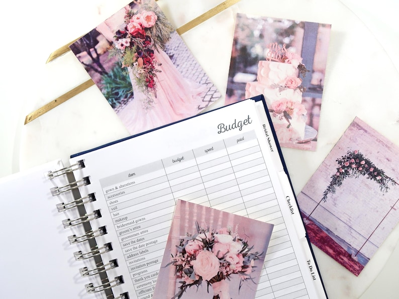 May include: A wedding budget planner with a checklist and to-do list. The planner has a white background with black text and lines. The planner is open to the budget page. There are several photos of wedding decorations on the pages of the planner.