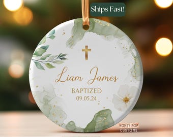 May include: Round white ceramic ornament with gold cross, green watercolor accents, and gold text. The ornament says "Liam James BAPTIZED 09.05.24". A gold ribbon is attached for hanging. The words "Ships Fast!" are in a green box.