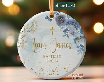 May include: Round ceramic ornament with a gold ribbon for hanging. The ornament features a watercolor design with blue floral accents, gold text that reads "Liam James," and the word "Baptized" with a date. Gold glitter dots are scattered across the bottom.