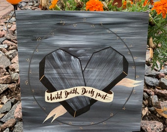 May include: Black wooden sign with a white painted heart shape made of two coffins. The coffins are tied together with a white ribbon that says "Until Death Do Us Part". The background is a dark gray with gold glitter.