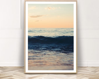 May include: A photograph of a blue ocean wave crashing on the shore at sunset. The sky is a soft orange and pink color, and the water is a deep blue.