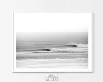 May include: A black and white photograph of waves crashing on a beach. The photo is taken from a distance, showing the waves rolling in and breaking on the shore. The sky is overcast and the water is a light grey.