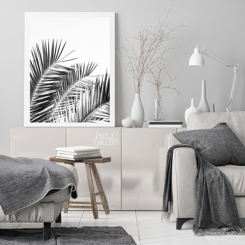 May include: Black and white photograph of palm tree leaves in a white frame. The frame is hanging on a white wall above a white cabinet with a wooden top. The cabinet is decorated with a vase of white branches, a white vase, and a white lamp.