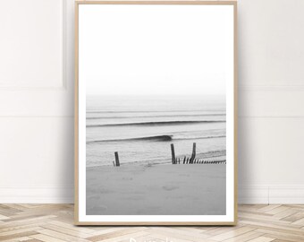 May include: Black and white photograph of a beach scene with waves crashing on the shore. A wooden fence is partially submerged in the sand.