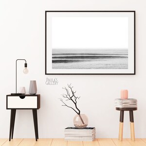 Op de afbeelding: Zwart-wit foto van oceaan golven ingelijst in een zwart frame hangend aan een witte muur. Een klein tafeltje met een lamp en decoratieve voorwerpen staat aan de linkerkant van de afbeelding. Een klein tafeltje met een stapel boeken en een vaas met een tak staat aan de rechterkant van de afbeelding.