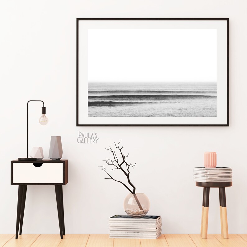 Op de afbeelding: Zwart-wit foto van oceaan golven ingelijst in een zwart frame hangend aan een witte muur. Een klein tafeltje met een lamp en decoratieve voorwerpen staat aan de linkerkant van de afbeelding. Een klein tafeltje met een stapel boeken en een vaas met een tak staat aan de rechterkant van de afbeelding.