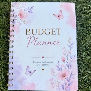 Op de afbeelding: Een spiraalgebonden budgetplanner met een witte kaft voorzien van aquarelachtige roze en paarse bloemenillustraties en twee vlinders. De tekst luidt BUDGET Planner en Organise tes finances avec sérénité, geaccentueerd door kleine hartjes.