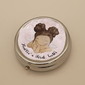 May include: A silver keepsake box with a pink and white design featuring a cartoon girl with brown hair and wings. The text "Hallie's First Teeth" is printed on the box.