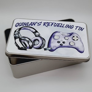 May include: A silver metal tin with a white lid. The lid features a purple and black illustration of a gaming headset and a video game controller. The text "QUINLAN'S REFUELLING TIN" is printed in purple on the lid.