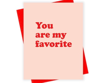 May include: A greeting card with the text "You are my favorite" in red, on a light pink background. The card is set against a red envelope, creating a contrasting color scheme. The design is simple and bold.