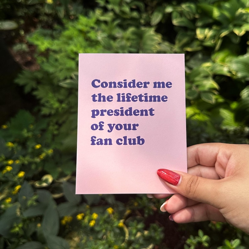May include: A rectangular light pink card featuring the dark purple text "Consider me the lifetime president of your fan club" printed in a bold, sans-serif font. The card is held against a background of green foliage and yellow flowers.