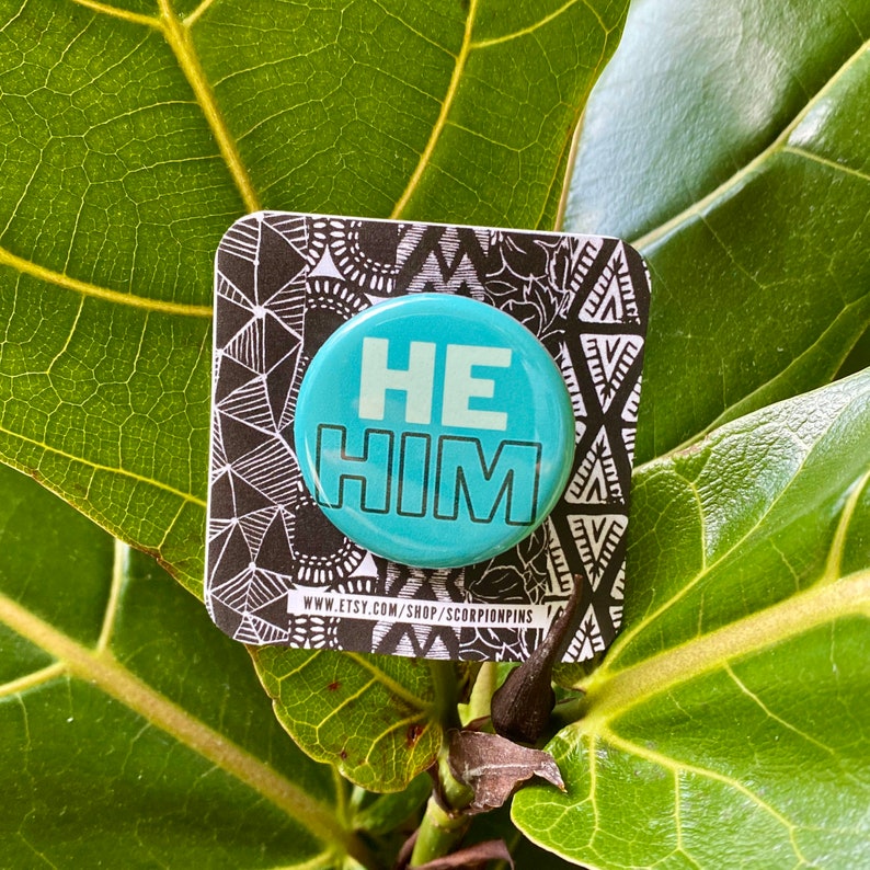 Pu&ograve; includere: Un bottone rotondo azzurro con le parole "HE HIM" in lettere bianche. Il bottone &egrave; su una carta a motivi bianchi e neri, su uno sfondo di foglie verdi. L'URL del negozio Etsy &egrave; visibile sulla carta.