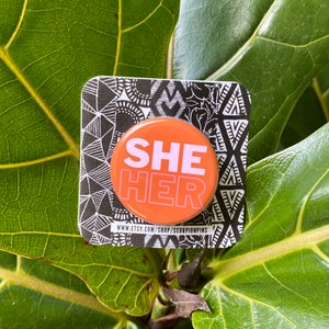 Pode incluir: Um botão redondo laranja com o texto "SHE HER" em branco e rosa. O botão está sobre um cartão com padrão preto e branco, em um fundo de folhas verdes. O URL da loja Etsy é visível na parte inferior.