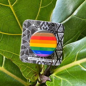 May include: A round button pin with a rainbow design, featuring horizontal stripes in brown, red, orange, yellow, green, blue, and purple. The pin is on a black and white patterned card with the text "www.etsy.com/shop/scorpionpin".
