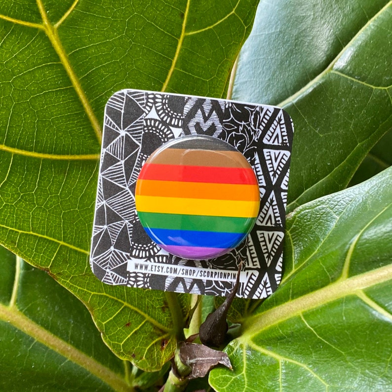 May include: A round button pin with a rainbow design, featuring horizontal stripes in brown, red, orange, yellow, green, blue, and purple. The pin is on a black and white patterned card with the text "www.etsy.com/shop/scorpionpin".