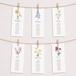 Può includere: Sei biglietti rettangolari bianchi con illustrazioni floreali e nomi, sospesi con mollette di legno su uno spago marrone. I fiori includono calendula, campanula, margherita, viola, ranuncolo e trifoglio. Ogni biglietto ha un elenco di nomi.