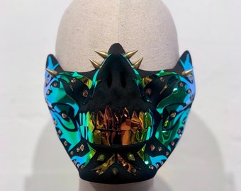 May include: A black mask with iridescent teal and gold accents, featuring a stylized skull design. The mask has gold spikes and studs, and is displayed on a mannequin head. The mask is a unique accessory.
