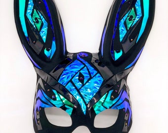 May include: A black bunny mask with long, upright ears. The mask features iridescent blue and green geometric shapes and accents. The edges are outlined in black, and small, decorative studs are placed around the mask.