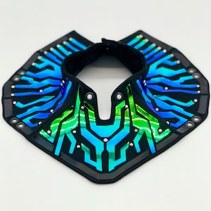 May include: A futuristic collar with a black fabric base, adorned with iridescent blue and green geometric shapes. The collar features small, sparkling embellishments and a hook-and-loop closure.