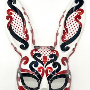 May include: A white rabbit mask with long ears, decorated with black and red swirling patterns. The mask features a red dotted mesh design and is suitable for costume parties or themed events.