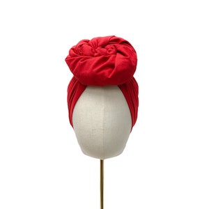 May include: A red turban head wrap with a knotted top, displayed on a white mannequin head.