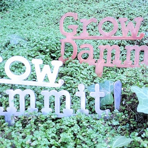 Puede incluir: Dos letreros de jard&iacute;n de metal con el texto "Grow Dammit!" en una fuente vintage. El letrero superior es de color cobre y el letrero inferior es plateado.