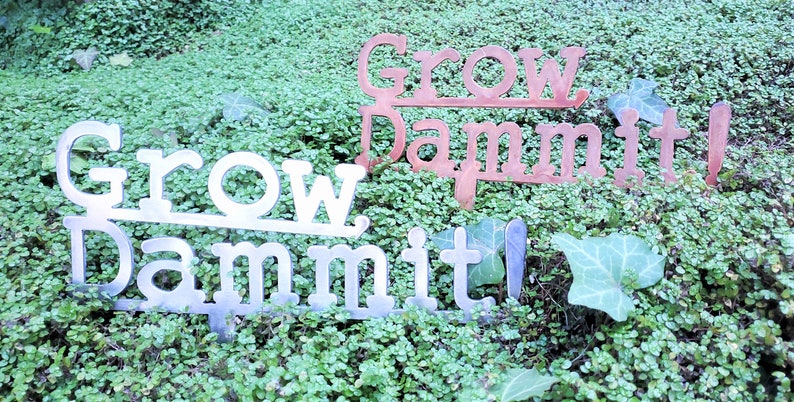 Puede incluir: Dos letreros de jard&iacute;n de metal con el texto "Grow Dammit!" en una fuente vintage. El letrero superior es de color cobre y el letrero inferior es plateado.