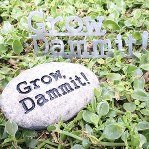 Puede incluir: Una piedra gris con el texto "Grow, Dammit!" grabado en ella. La piedra est&aacute; rodeada de follaje verde. Un letrero de metal con el texto "Grow, Dammit!" es visible en el fondo.