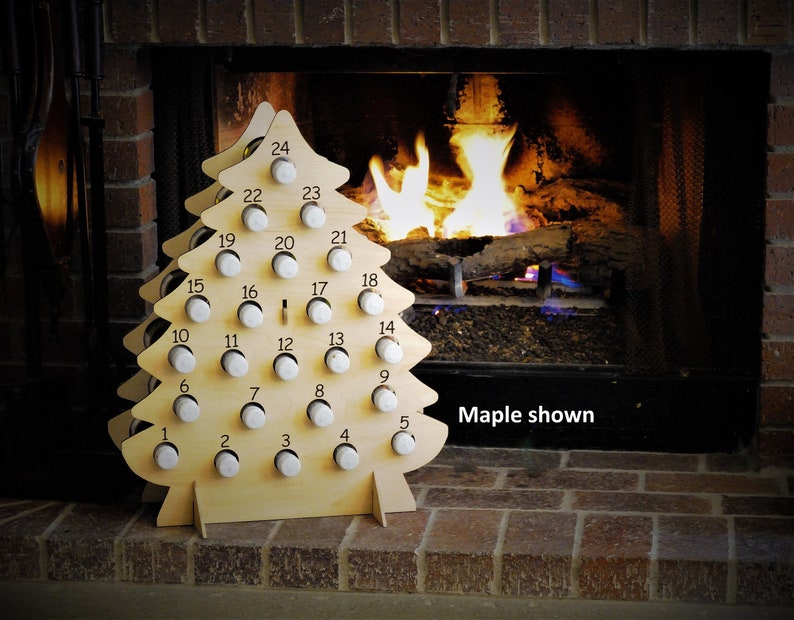 May include: A wooden Christmas tree advent calendar with 24 numbered slots for holding small gifts or treats. The tree is made of light wood and has a natural finish. The calendar is shown in front of a fireplace with a fire burning inside.