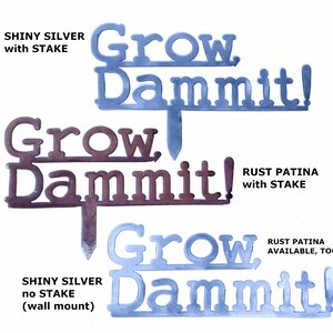 Puede incluir: Dos estacas de jard&iacute;n de metal con el texto "Grow Dammit!" en un estilo r&uacute;stico. La estaca superior es de plata brillante con una estaca, la estaca inferior es de color marr&oacute;n oxidado con una estaca. La estaca inferior tambi&eacute;n tiene una versi&oacute;n sin estaca para montaje en pared.