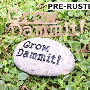Puede incluir: Una piedra gris con las palabras "Grow Dammit!" grabadas en ella. La piedra est&aacute; sentada en un lecho de hierba verde. Un letrero de metal con las palabras "Grow Dammit!" est&aacute; sentado en la hierba detr&aacute;s de la piedra.