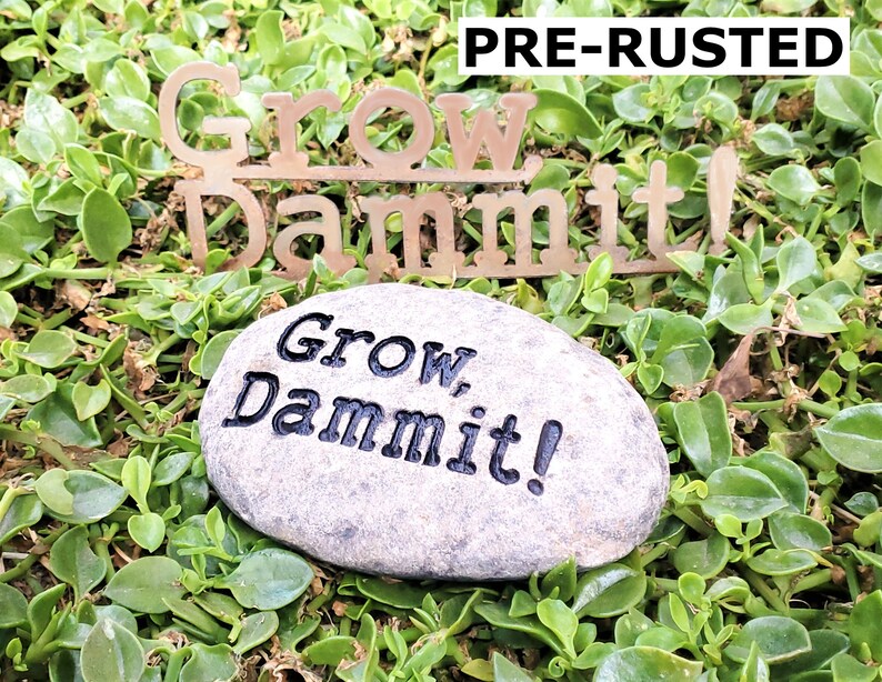 Puede incluir: Una piedra gris con las palabras "Grow Dammit!" grabadas en ella. La piedra est&aacute; sentada en un lecho de hierba verde. Un letrero de metal con las palabras "Grow Dammit!" est&aacute; sentado en la hierba detr&aacute;s de la piedra.