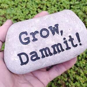 Puede incluir: Una piedra gris con las palabras "Grow, Dammit!" pintadas en negro.