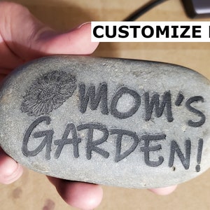 Puede incluir: Una piedra gris con las palabras "MOM'S GARDEN!" grabadas en ella. Tambi&eacute;n hay un peque&ntilde;o dise&ntilde;o de flor grabado en la piedra.