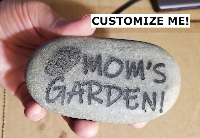 Puede incluir: Una piedra gris con las palabras "MOM'S GARDEN!" grabadas en ella. Tambi&eacute;n hay un peque&ntilde;o dise&ntilde;o de flor grabado en la piedra.