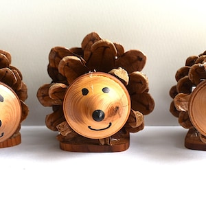 May include: Three hedgehog figurines crafted from brown pinecones with smooth, light-coloured wooden faces. Each figure features a painted black nose, two small black eyes, and a simple curved black line for a mouth, all mounted on a small wooden base.